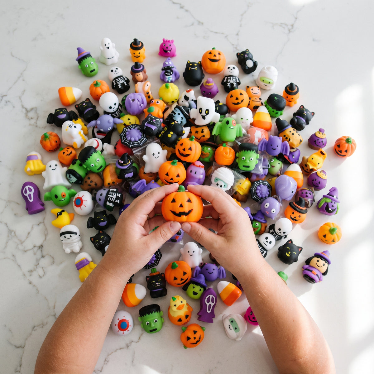 Dozens of mini Halloween mochi squishies spread across a white counter, a child's hands lifting an orange jack-o'-lantern from the middle