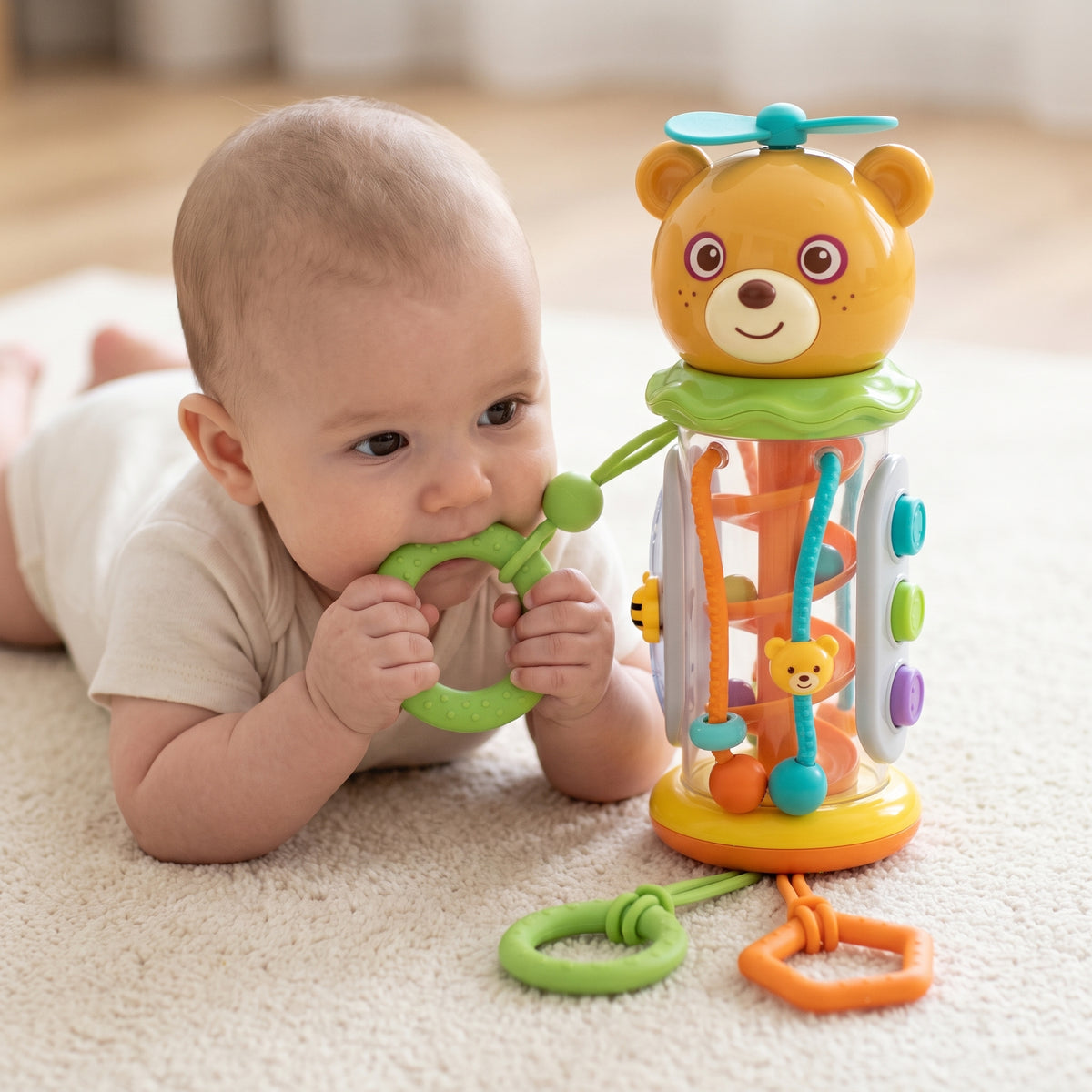 Baby on their tummy on a cream rug, holding the green silicone teething ring to their mouth while the bear rattle stands beside them