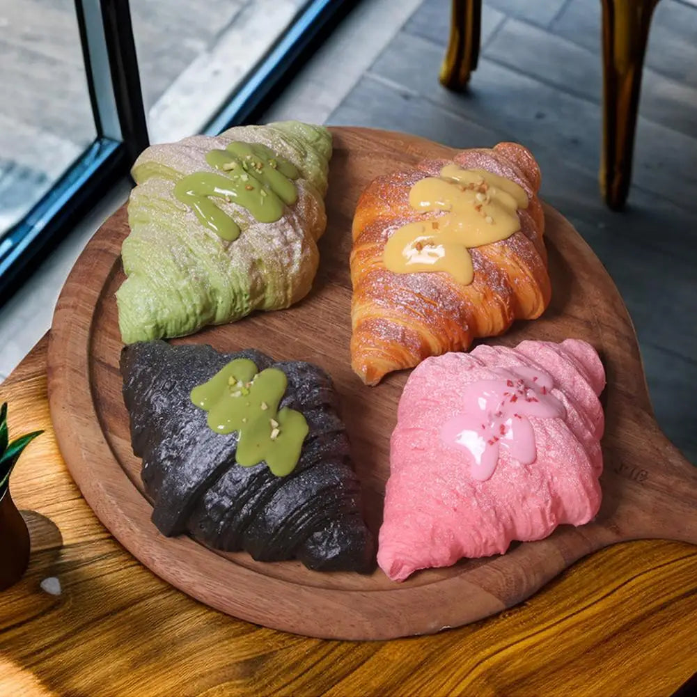 Four croissant squishies, golden, pink, green and black, on a wooden board