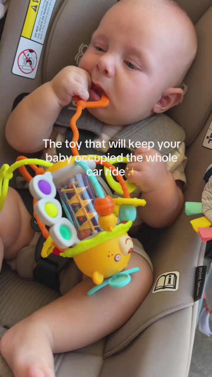 Baby strapped in a car seat chewing on the orange silicone ring of the bear rattle