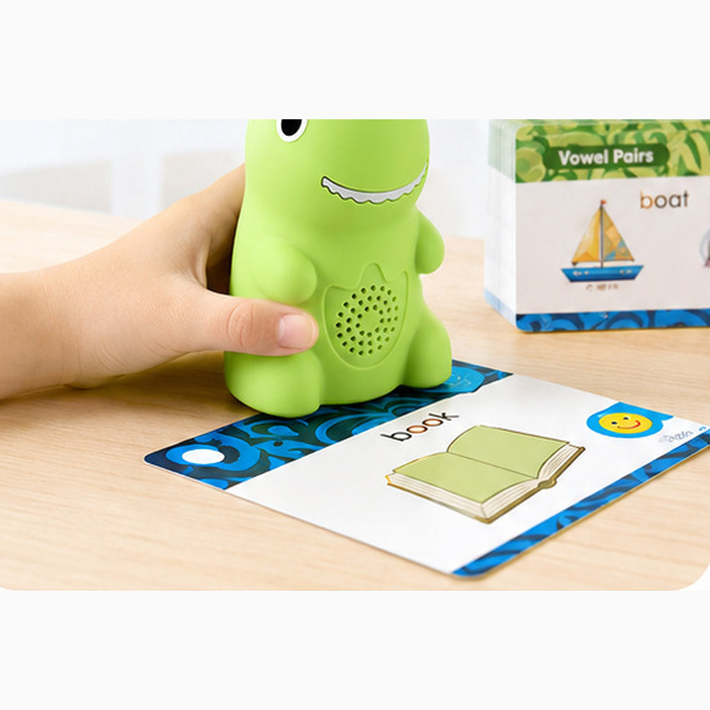 A child's hand rests the dinosaur reader on a phonics card showing the word book, with the vowel pairs card set standing behind